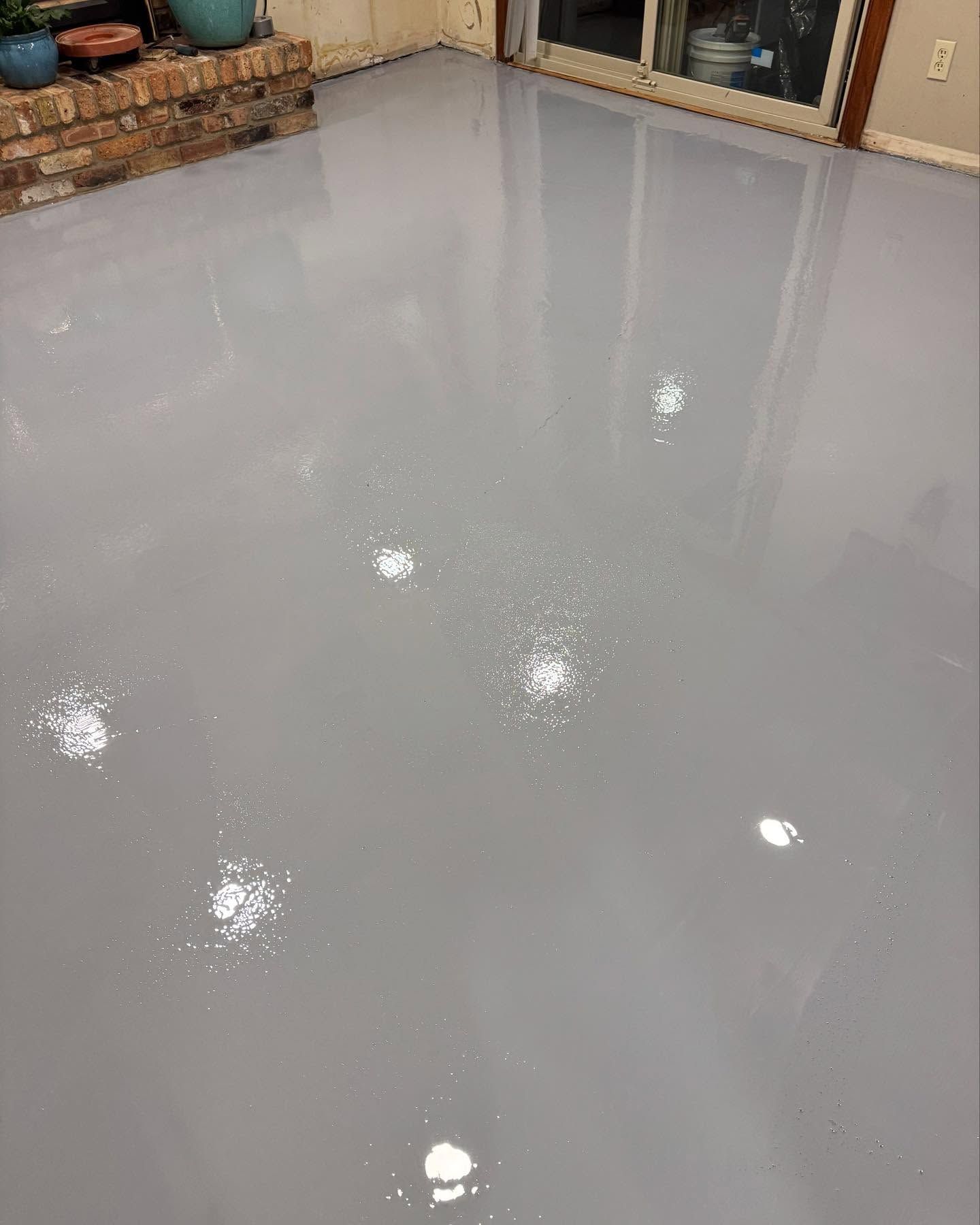 A smooth, glossy, light gray epoxy floor reflecting overhead lights in a room with a brick fireplace and glass doors.