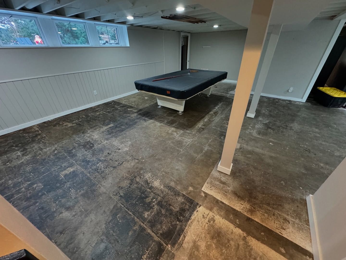 A basement with a pool table on a bare concrete floor, featuring gray walls, white wainscoting, and a support column.