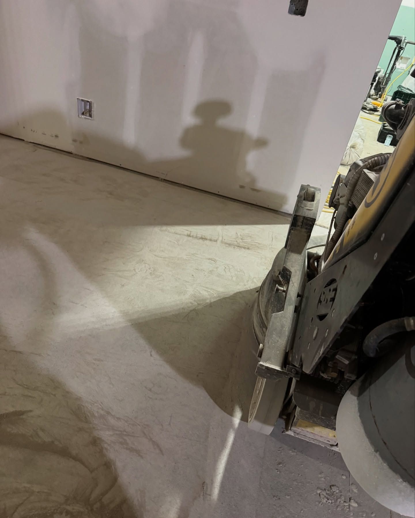 A close-up view of a floor sander in operation on a dusty concrete floor inside a room under construction.