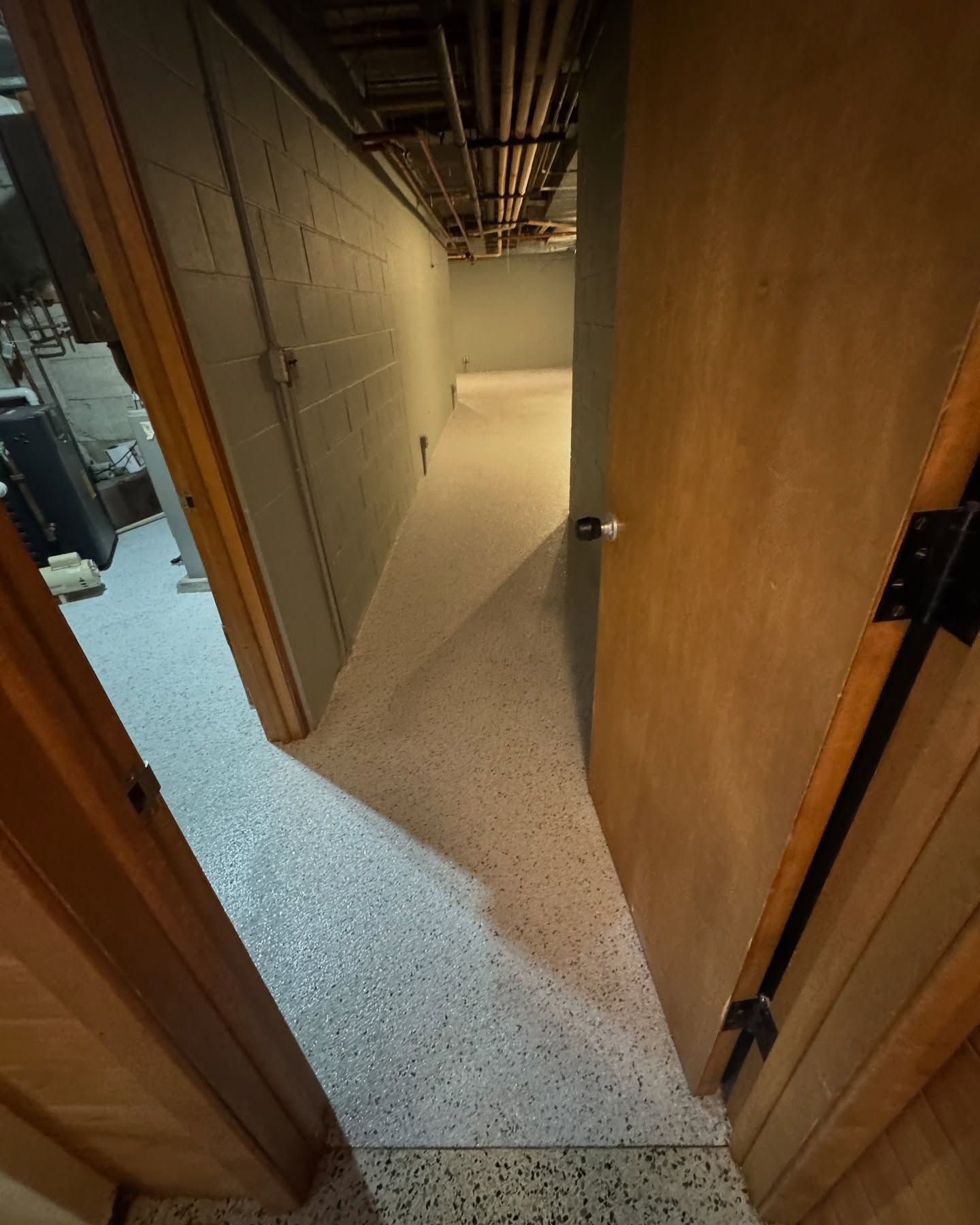A view through an open doorway looking into a basement corridor with light-colored, speckled flooring and grey walls.