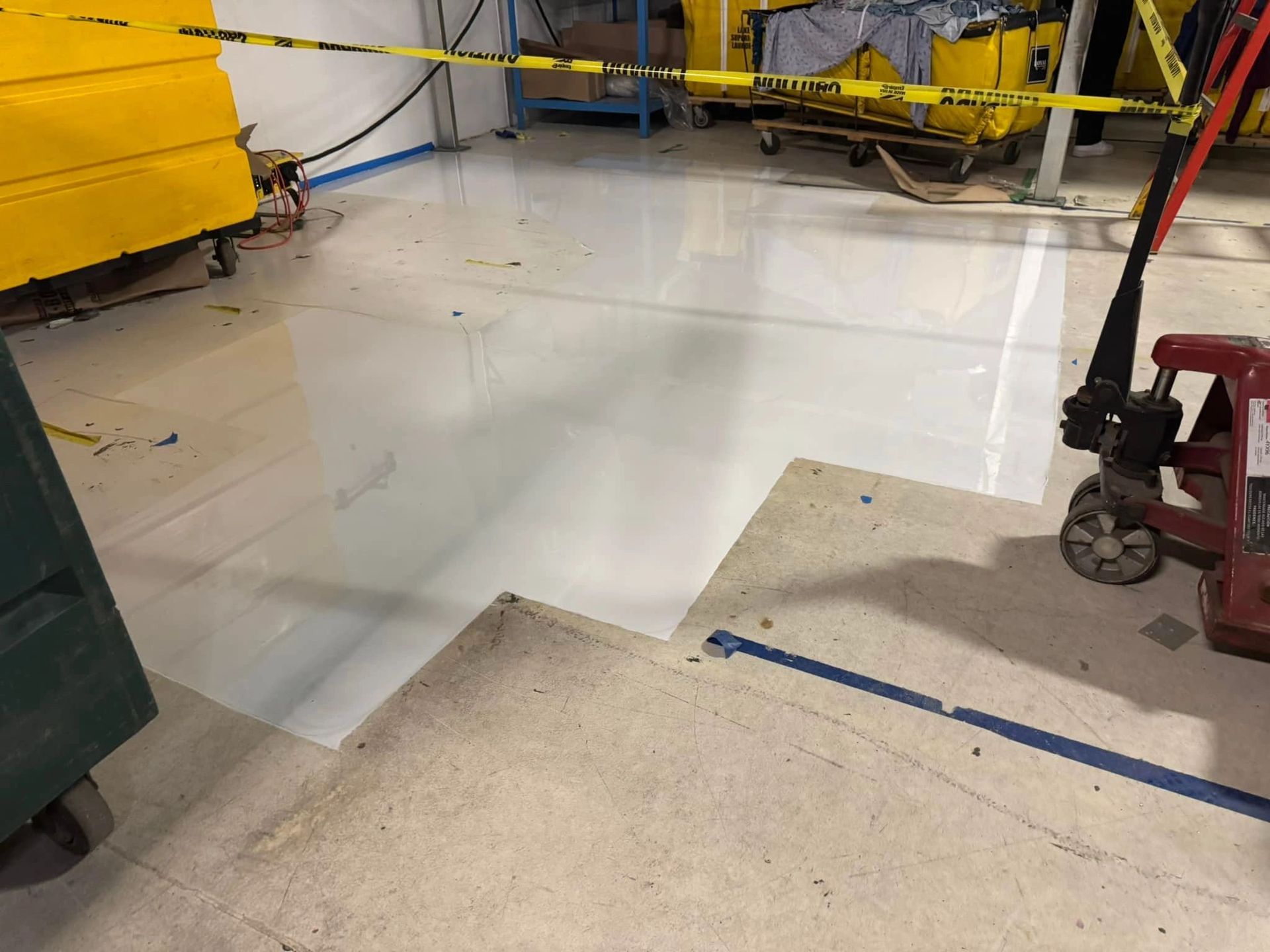 A shiny white epoxy floor patch in an industrial warehouse, bordered by a rough concrete floor and safety tape.