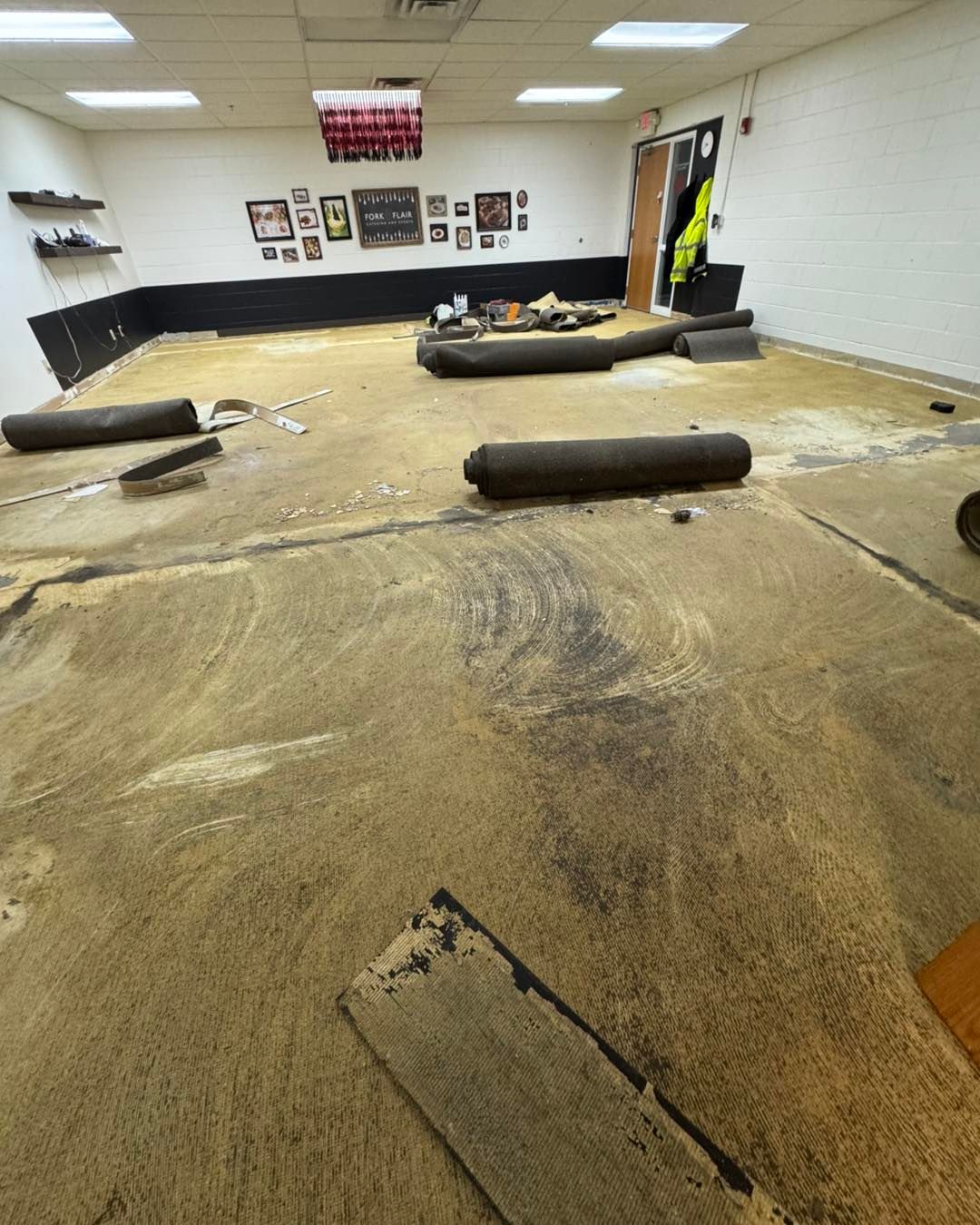 An empty room with bare concrete floors, rolled-up carpet remnants, and framed photos on the far wall.