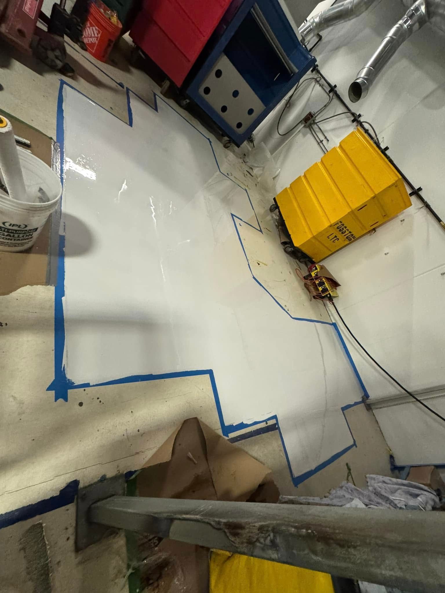 A bright white patch of fresh epoxy on a floor, outlined with blue painter’s tape inside an industrial workspace.