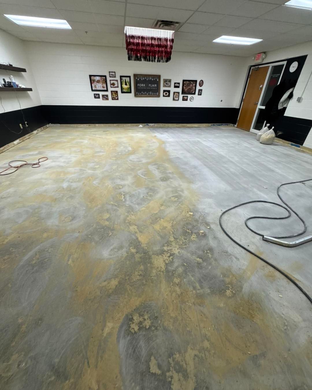 An empty room with a concrete floor half-sanded, a gallery wall with various framed pictures, and black lower wall trim.
