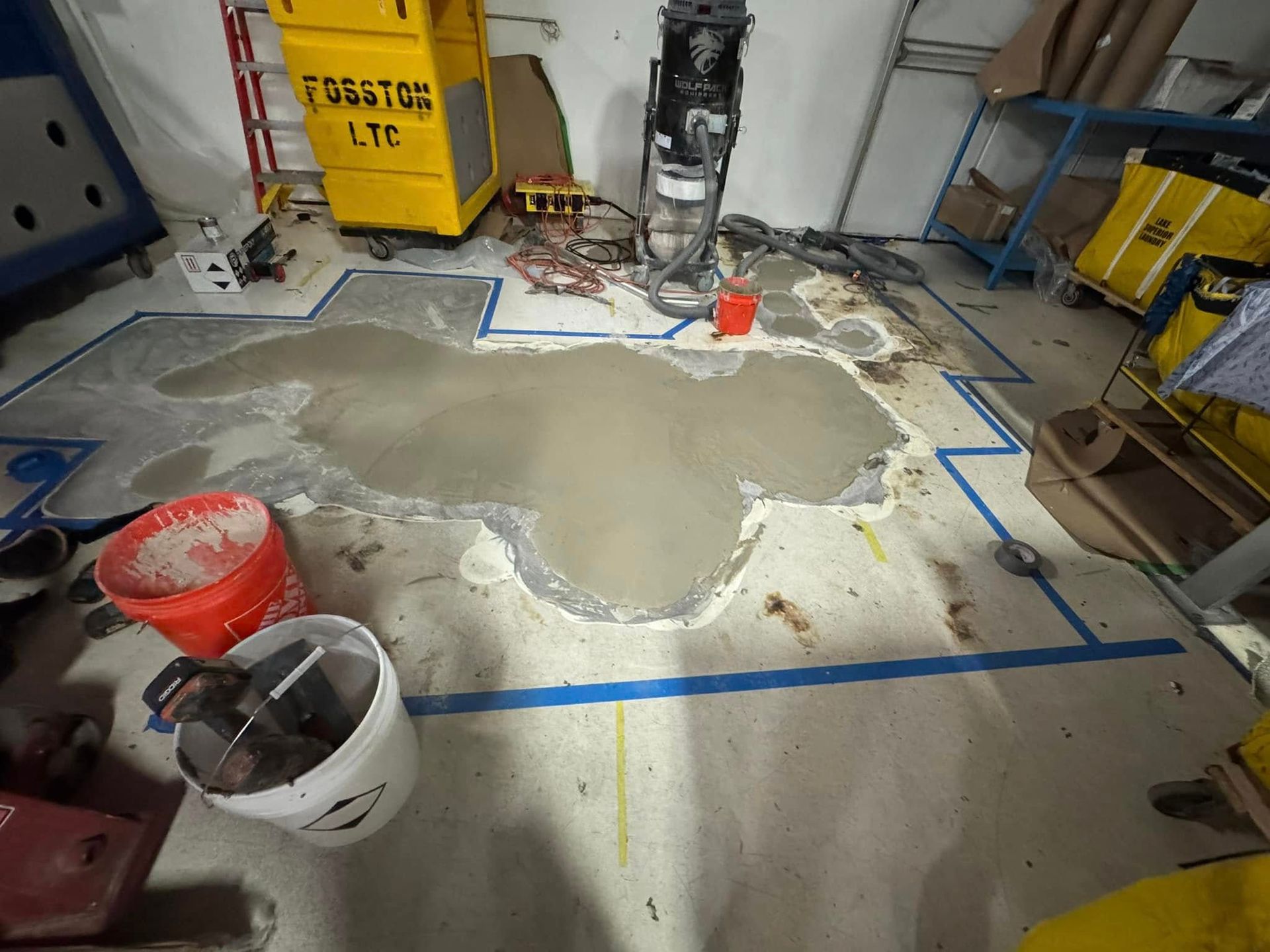 Wet concrete patch on a warehouse floor, surrounded by blue painter's tape, cleaning tools, and yellow storage bins.