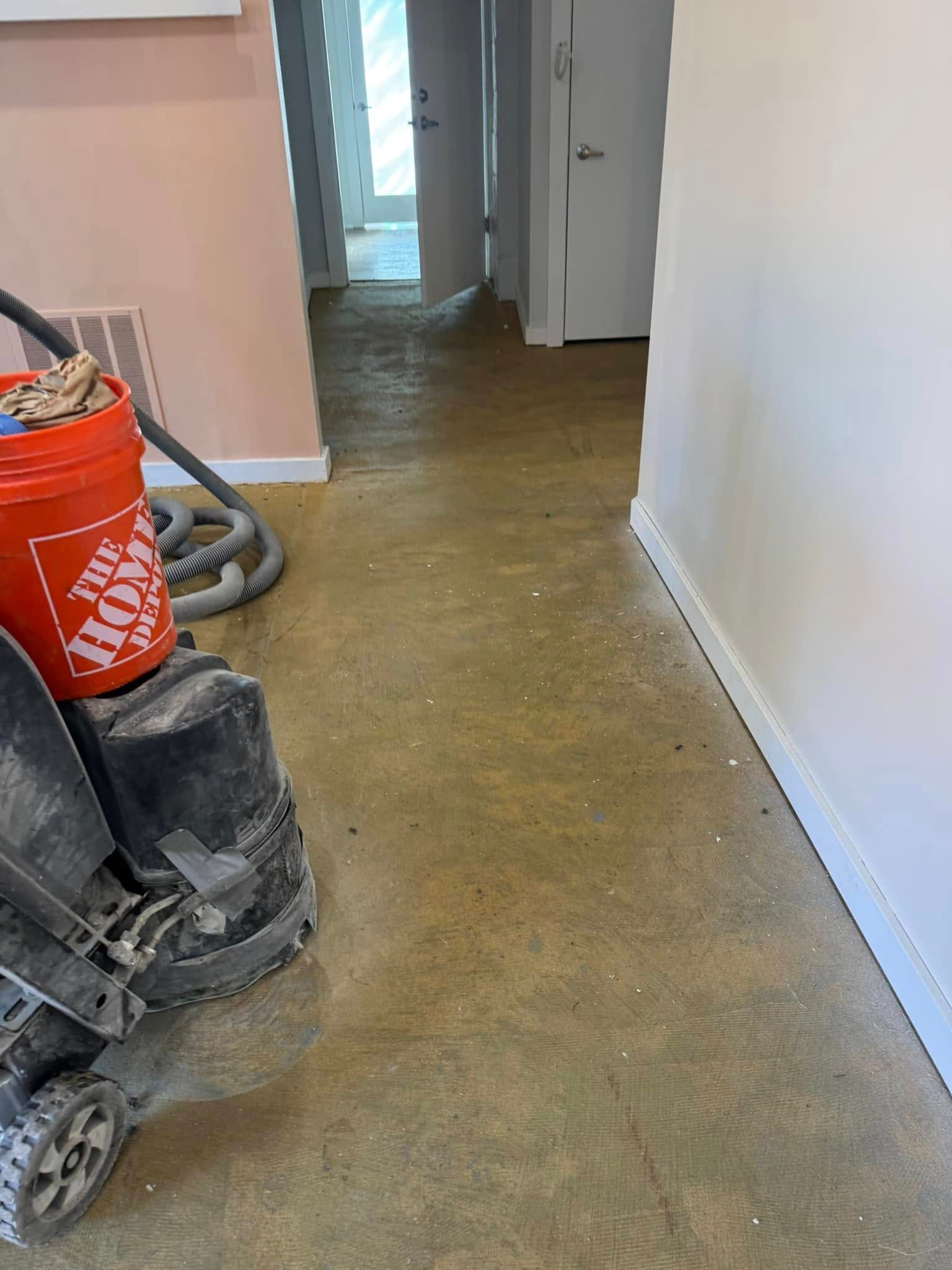 A floor sander and an orange Home Depot bucket sit on a dusty concrete floor in a room with white walls and a hallway.