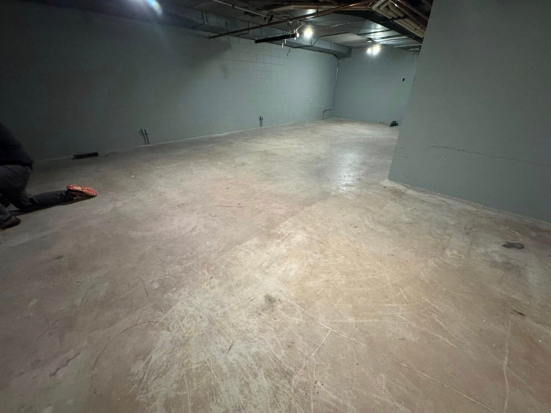 A person kneels on the concrete floor of a large, empty basement room with gray walls and an exposed ceiling.