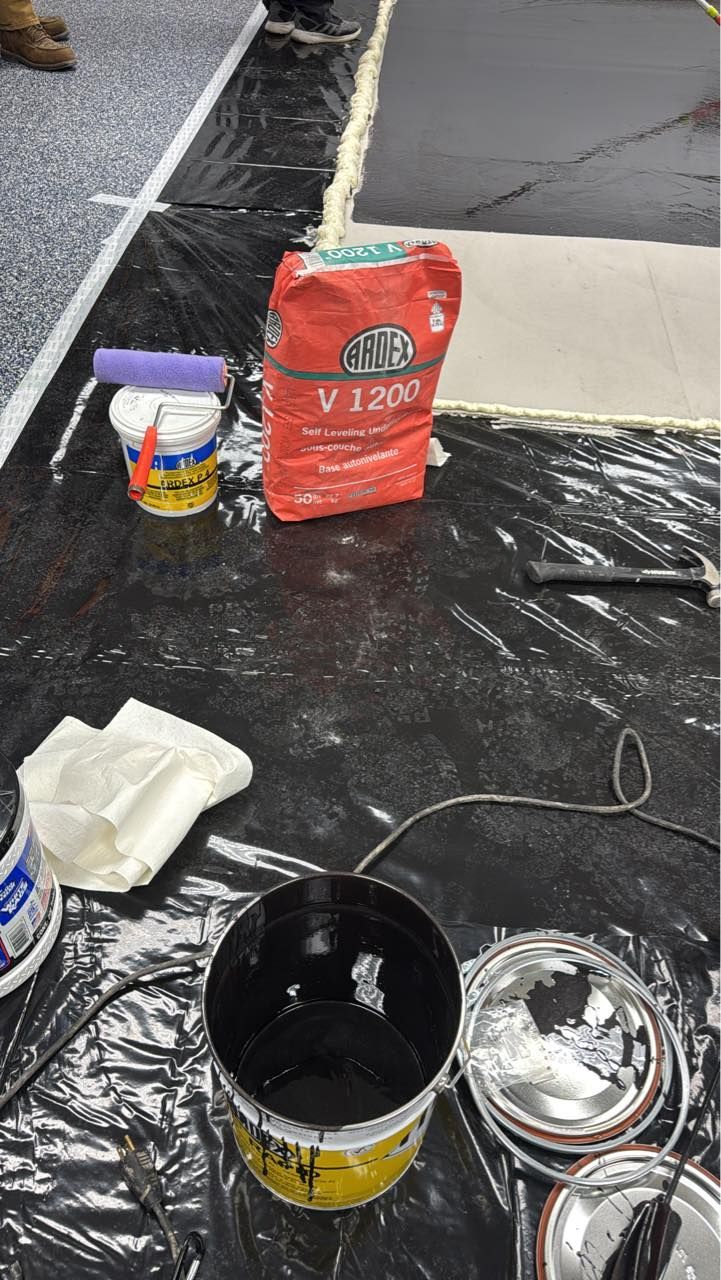 Construction supplies including a bag of V1200 grout, a paint bucket, and rollers on a plastic-covered floor.