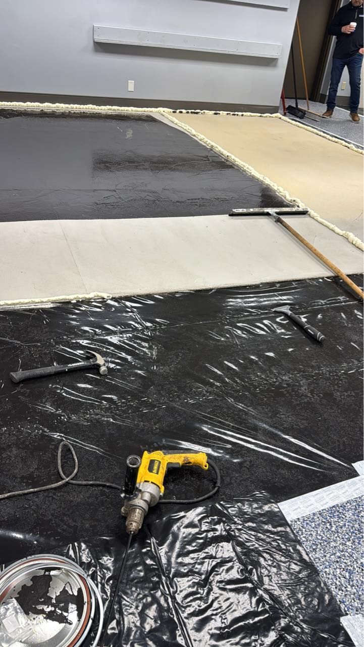 An indoor space with a floor under construction, showing grey and tan sections, a squeegee, tools, and a drill on plastic.