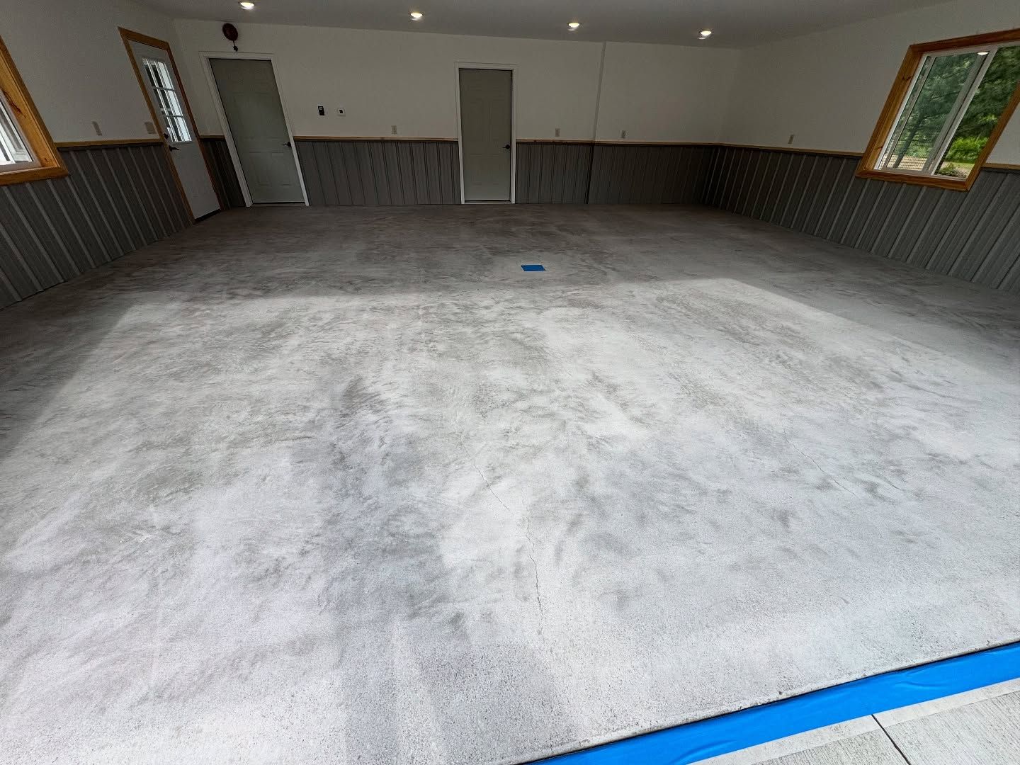 A large, empty garage featuring a mottled gray concrete floor, neutral wainscoting, two doors, and a window.