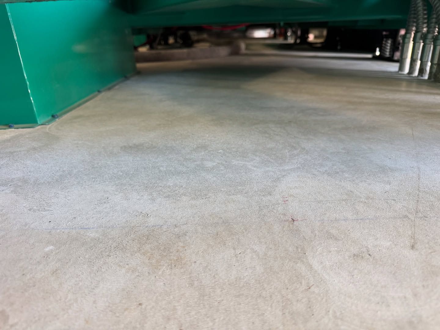 A view from floor level under a large green industrial machine, showing a smooth concrete floor and metal conduits.