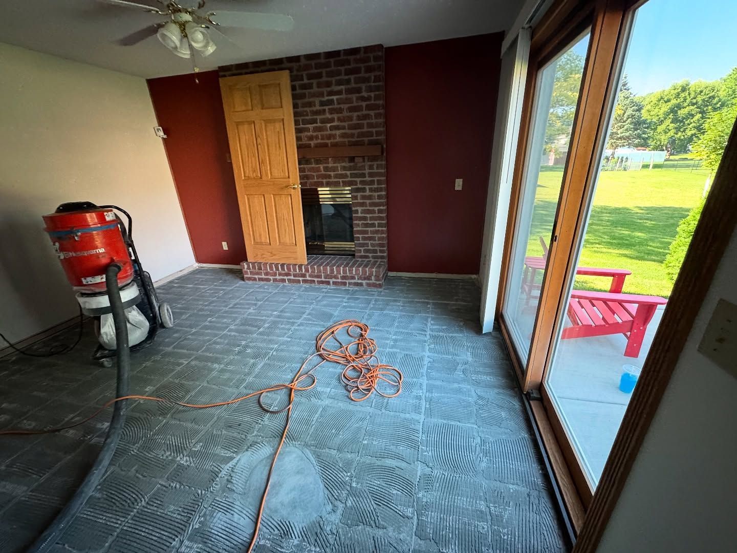 Interior room undergoing floor renovation with a red shop vacuum, exposed brick fireplace, and sliding glass patio doors.