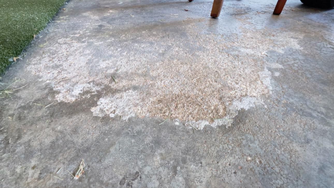 A patch of worn, weathered concrete flooring shows exposed aggregate near a patch of grass and chair legs.