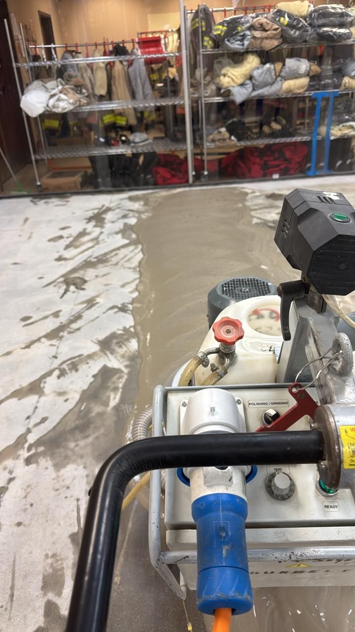 A floor grinding machine processes a concrete surface inside a storage room with shelves of gear in the background.