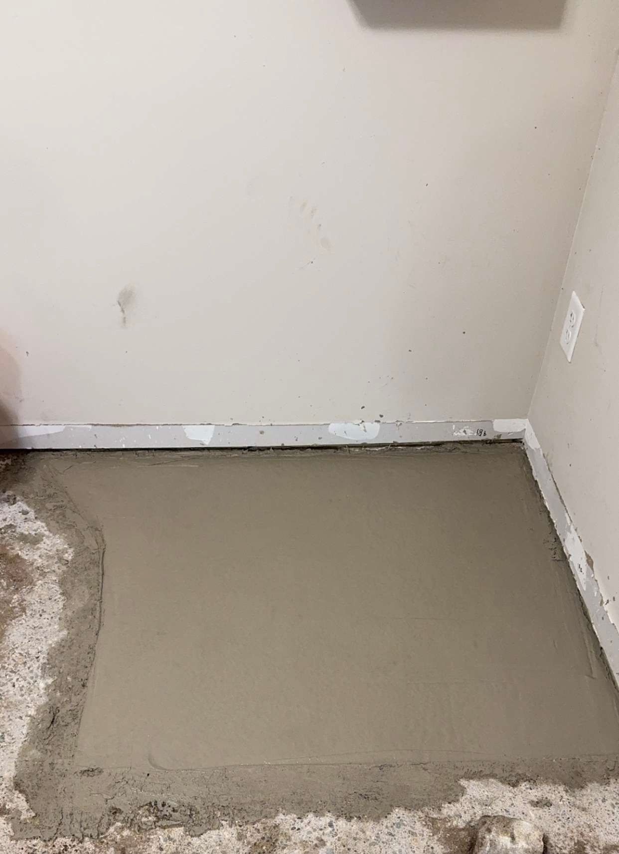 A patch of wet, gray cement filling a rectangular hole in a concrete floor against a beige wall.