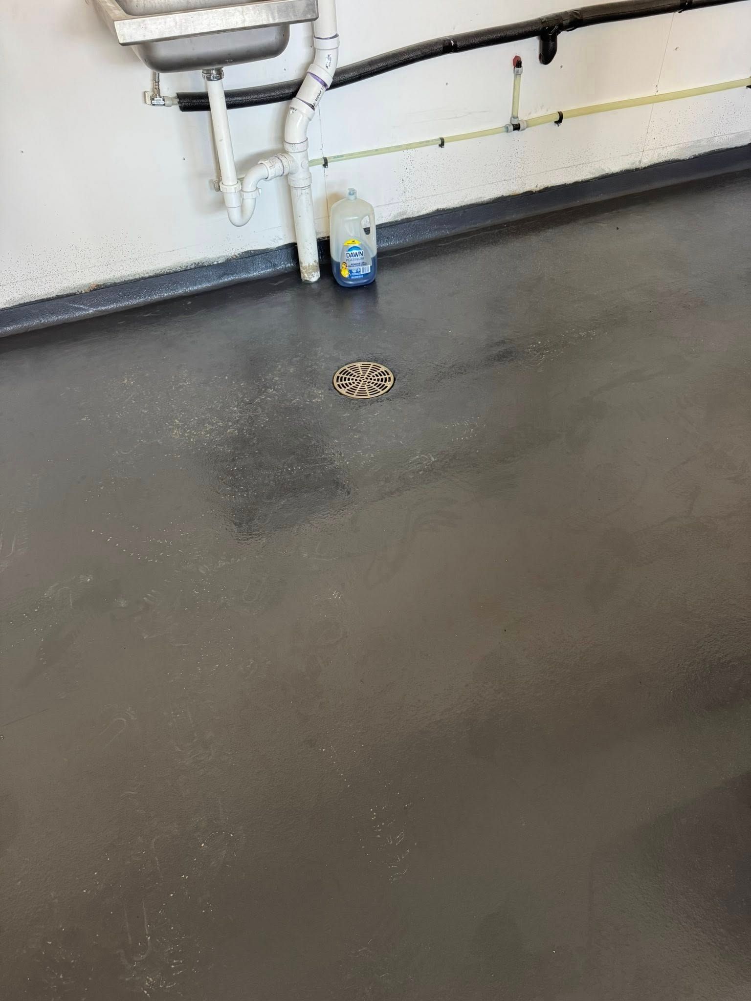 A gray floor with a circular drain, a utility sink pipe, and a white plastic jug against a white wall.