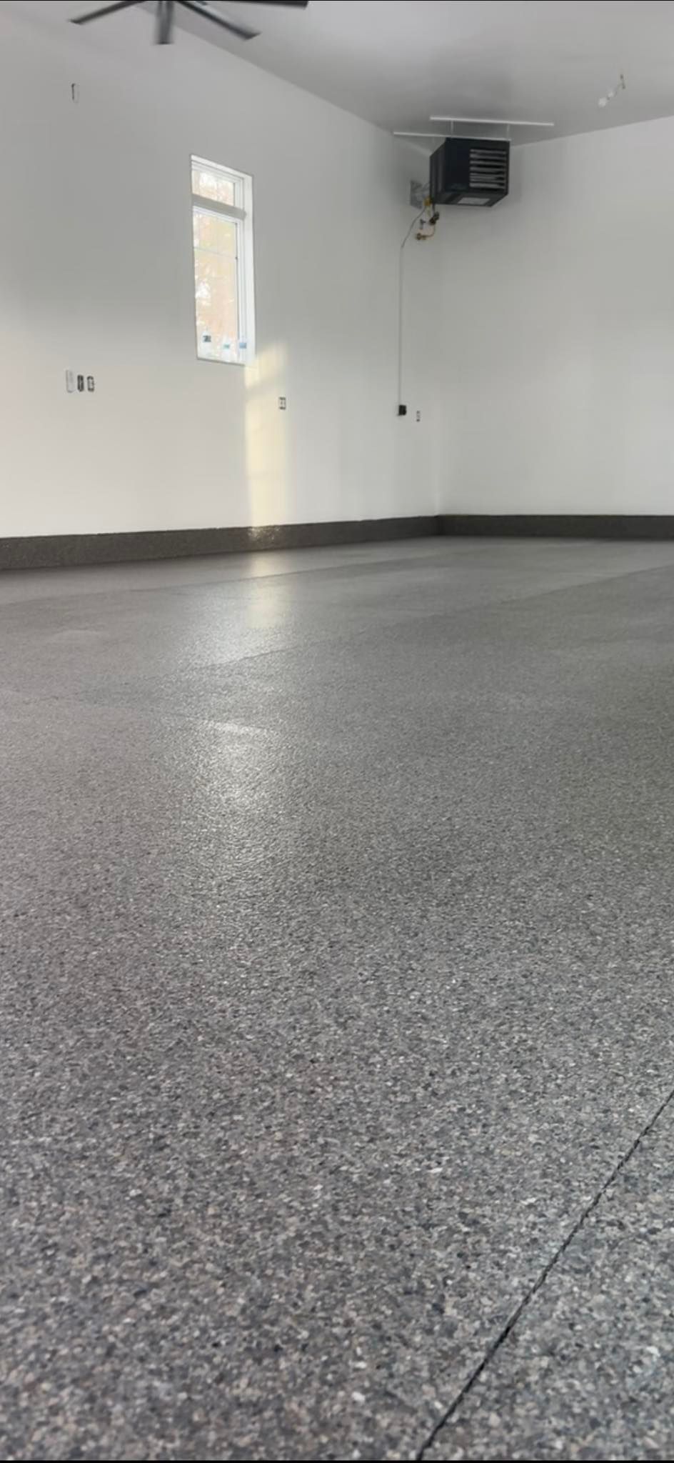 An empty garage with white walls, a gray speckled epoxy floor, a side window, and a ceiling-mounted heater.