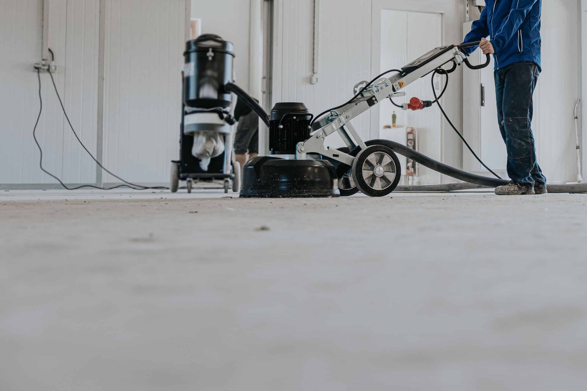 A person operating a heavy-duty floor grinder machine on a concrete surface in an unfinished room.