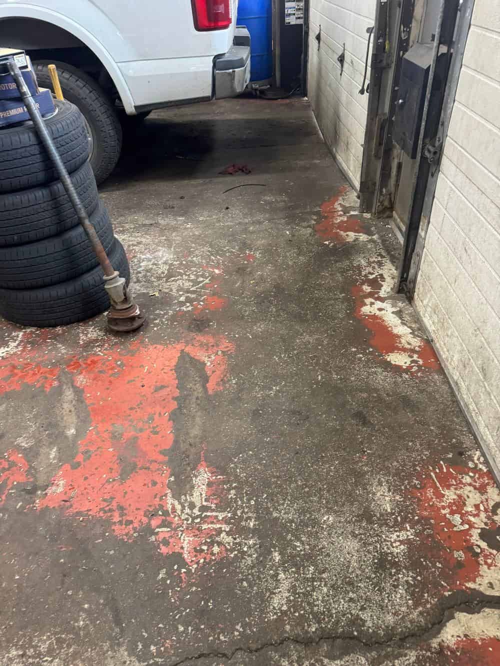 A stack of tires and a metal axle component on a worn, paint-chipped concrete floor in a garage near a white truck.