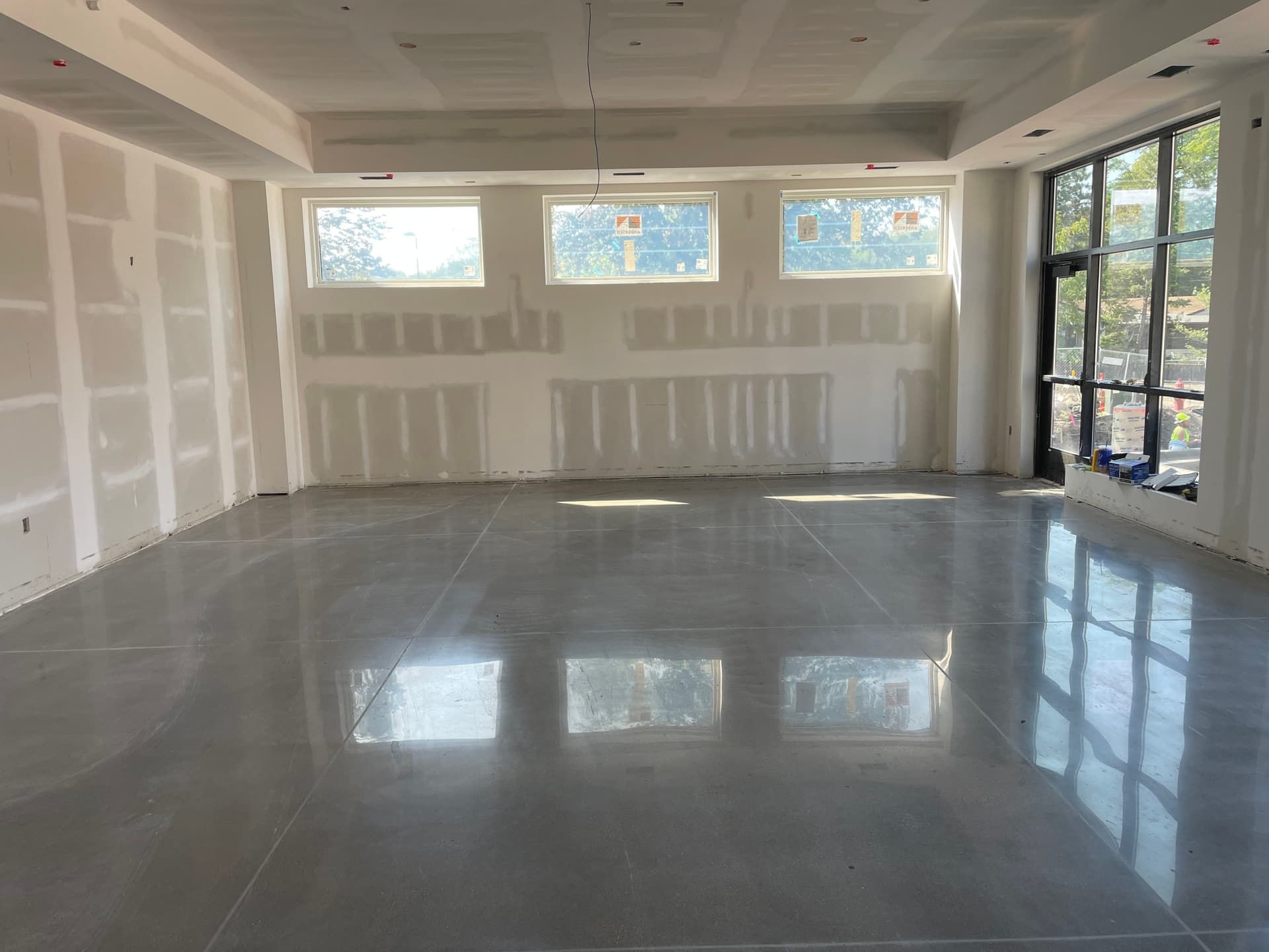 A wide, empty room under construction with finished gray concrete flooring, drywall walls, and large glass windows.