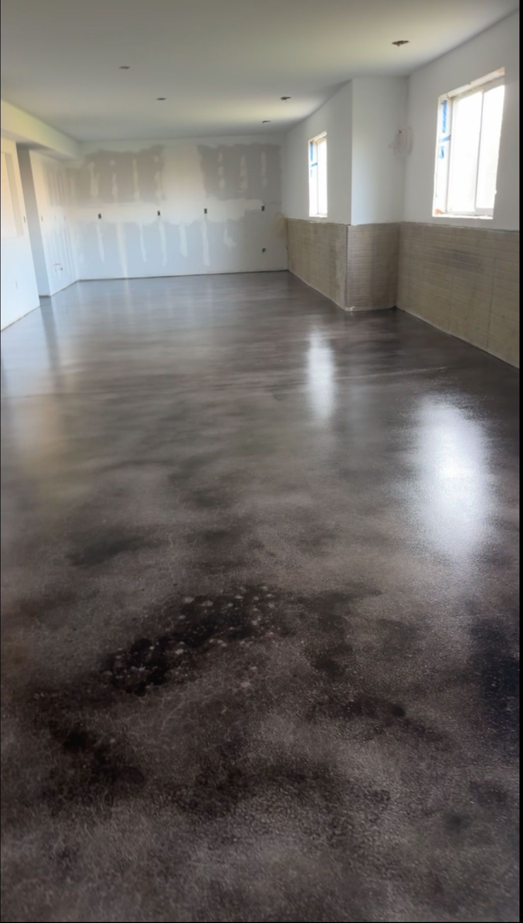 A long, empty room with freshly polished, mottled dark gray concrete floors and white walls with patches of dry plaster.