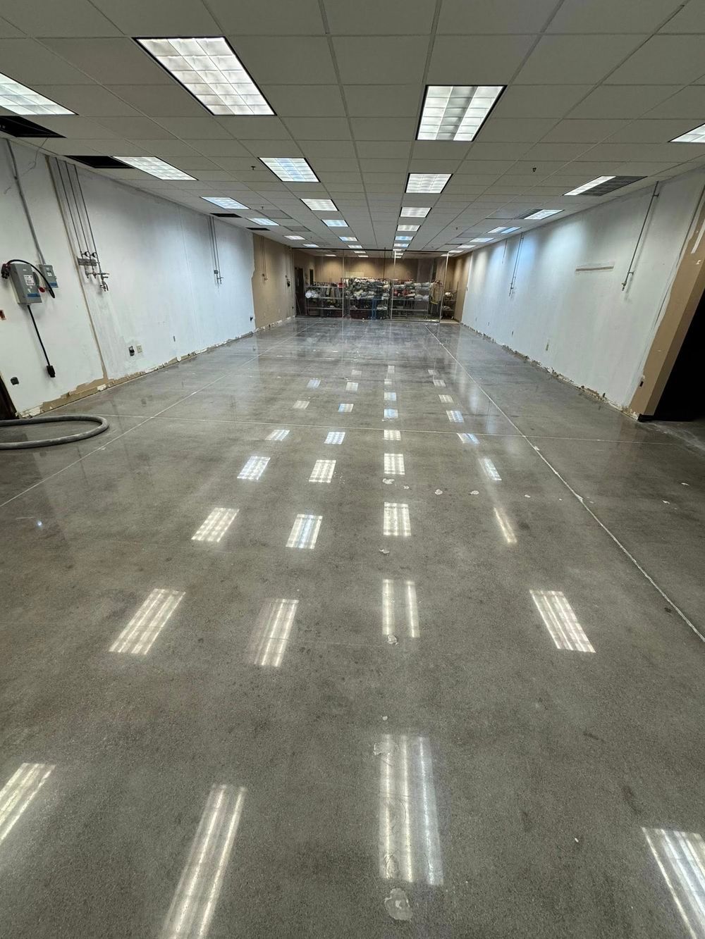 A wide, empty commercial space with polished concrete floors and drop ceiling lights, extending toward the back wall.
