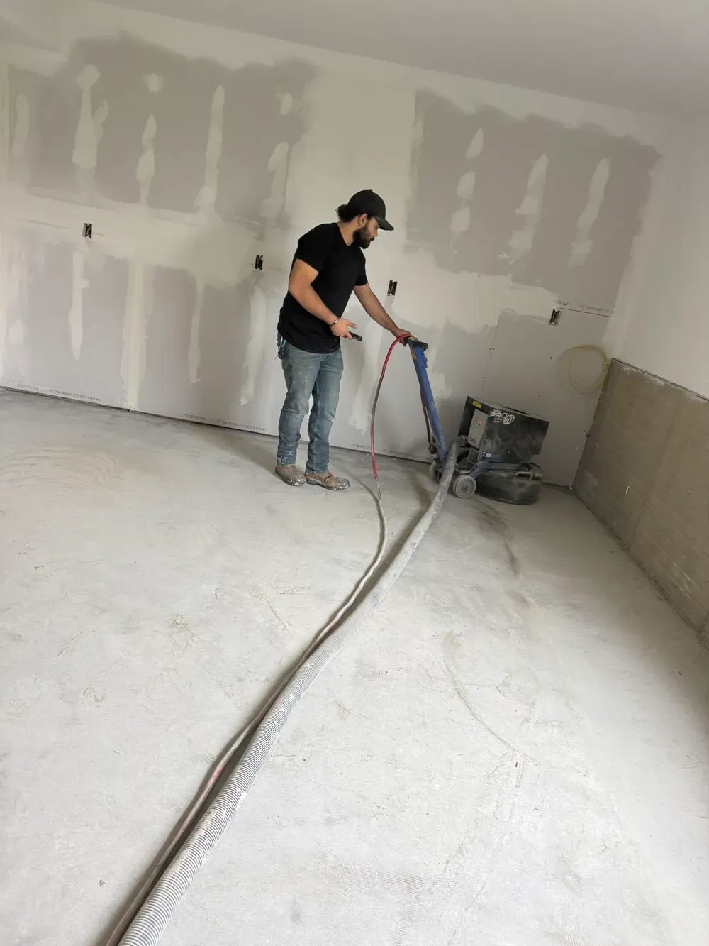 A person uses a floor sander in a room with unfinished drywall walls and a concrete floor.