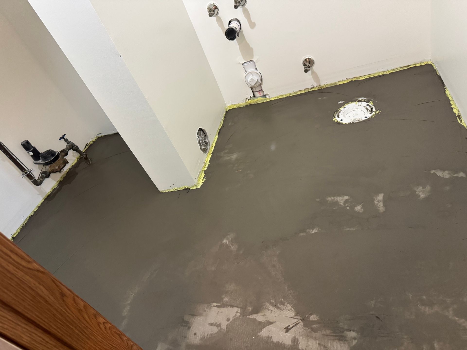 A small room with freshly poured gray floor leveling compound and visible wall plumbing pipes, set against white walls.
