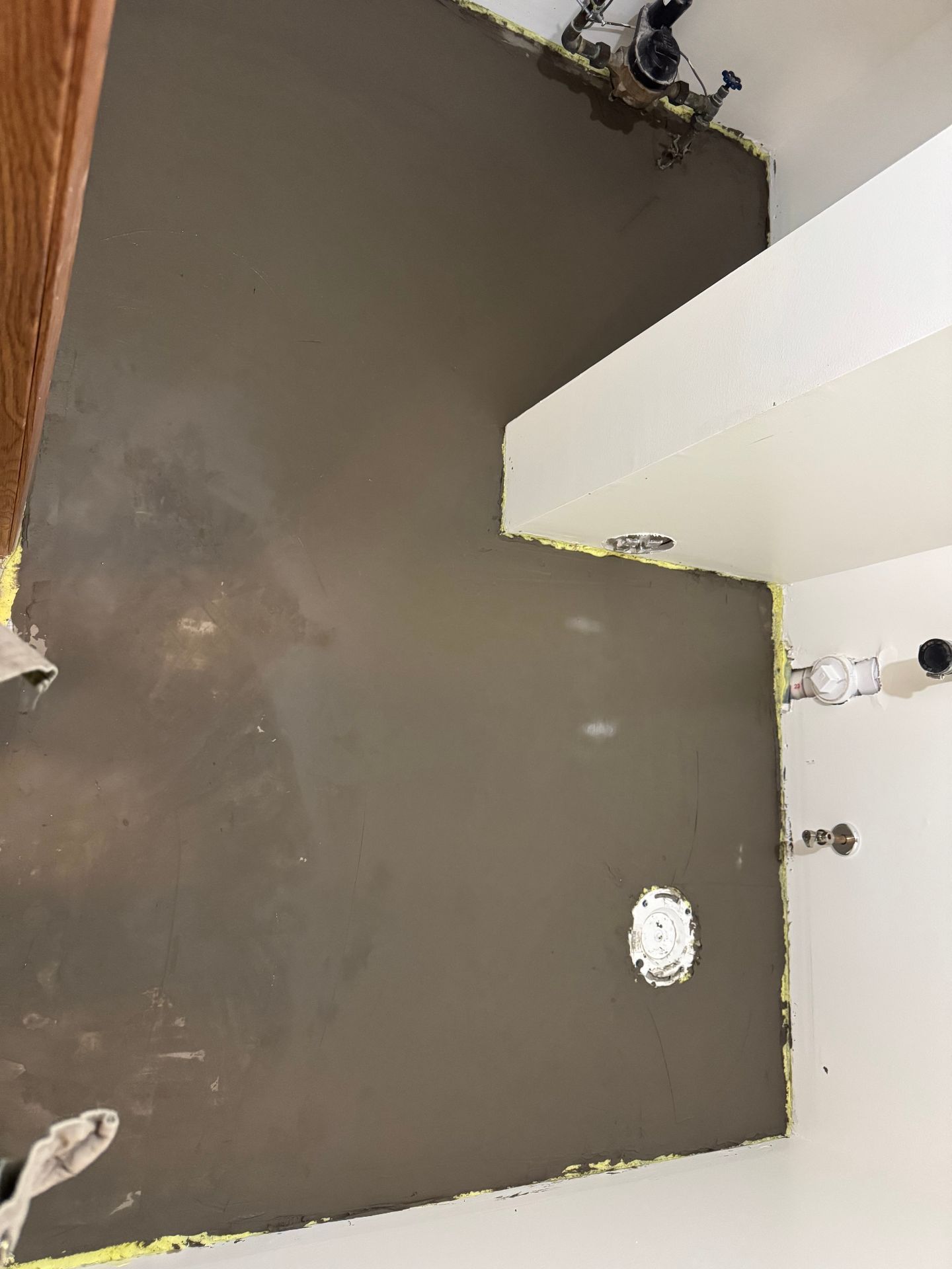 A freshly leveled concrete subfloor in a bathroom with plumbing pipes visible at the wall and a floor drain flange.