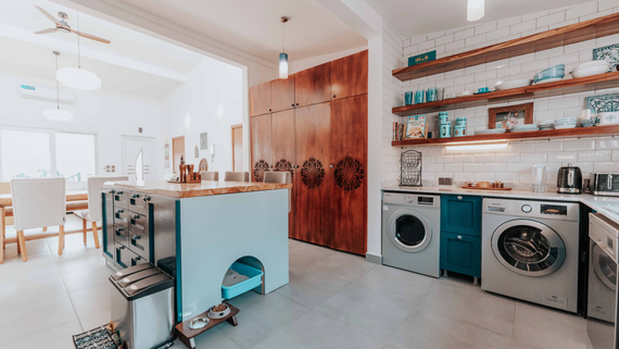 Bright open kitchen-laundry space with wooden shelves, washing machines, island, and dining area