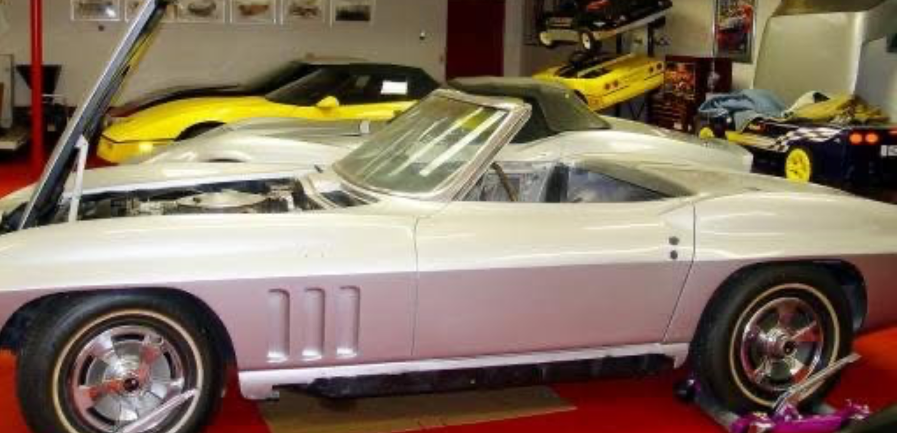 Silver and white classic convertible sports car displayed in a showroom, with red wheels and yellow cars behind it Silver and white classic convertible sports car displayed in a showroom, with red wheels and yellow cars behind it