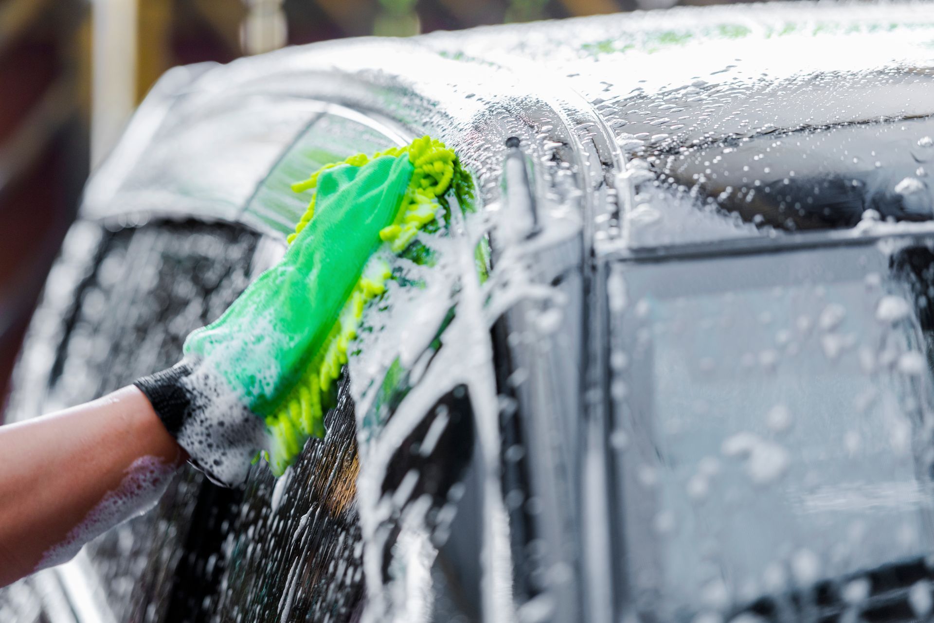 Hand washing a black car with a green sponge, soap suds and water splashing on the window