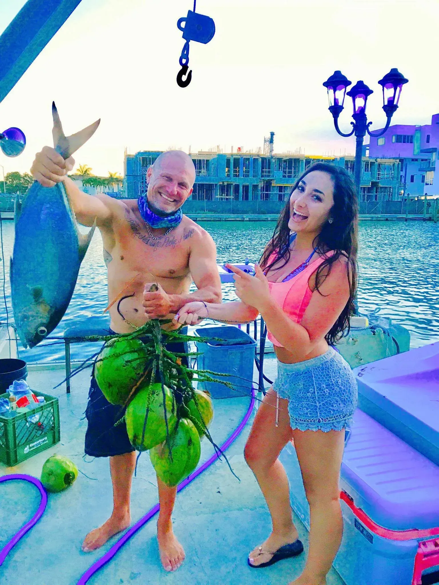 A man and a woman are standing on a dock holding a fish and coconuts.
