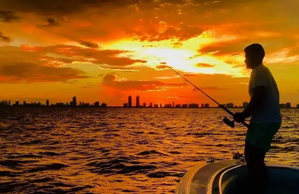 A man is fishing from a boat at sunset.