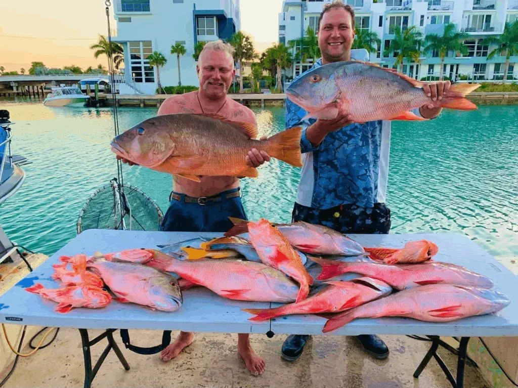 Two men are holding large fish in front of a table full of fish.