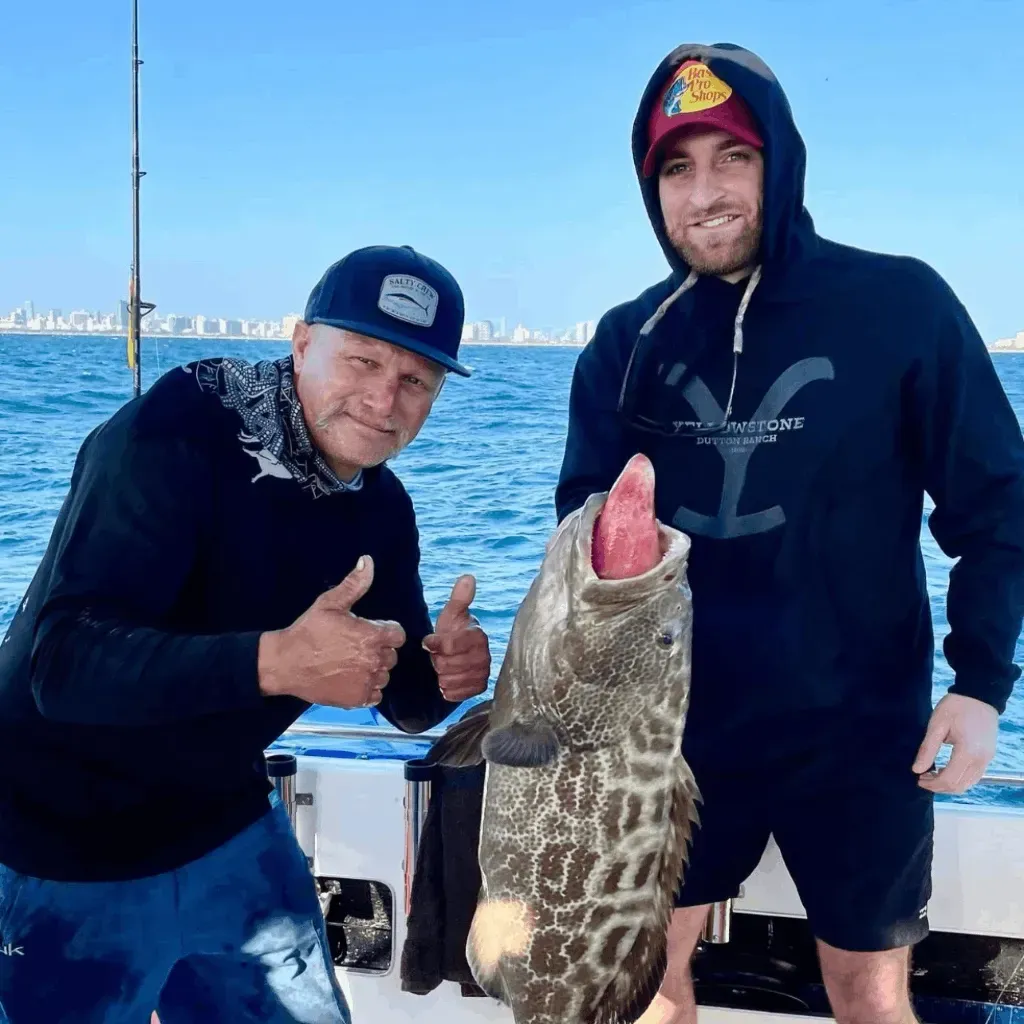 Two men are standing next to each other on a boat holding a large fish.