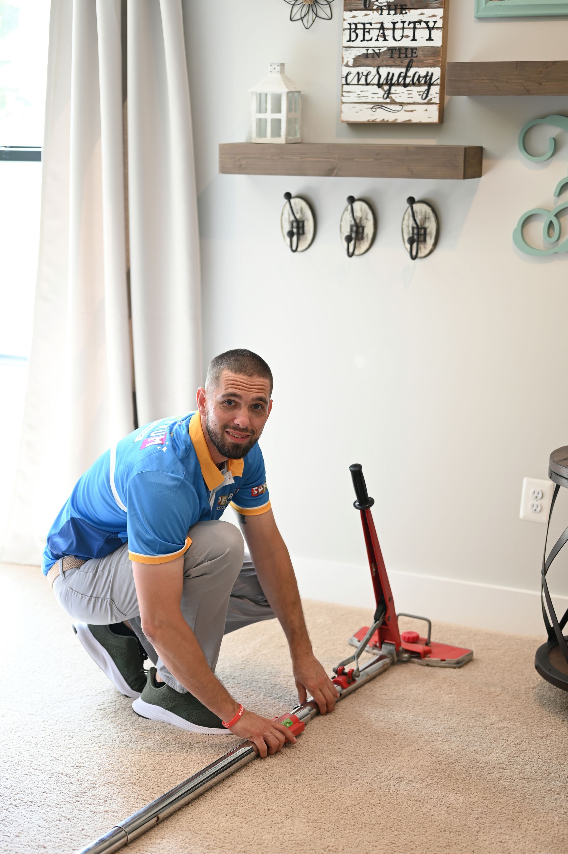 Man kneeling on carpet, assembling red carpet stretcher, smiling, in front of a wall with decor.