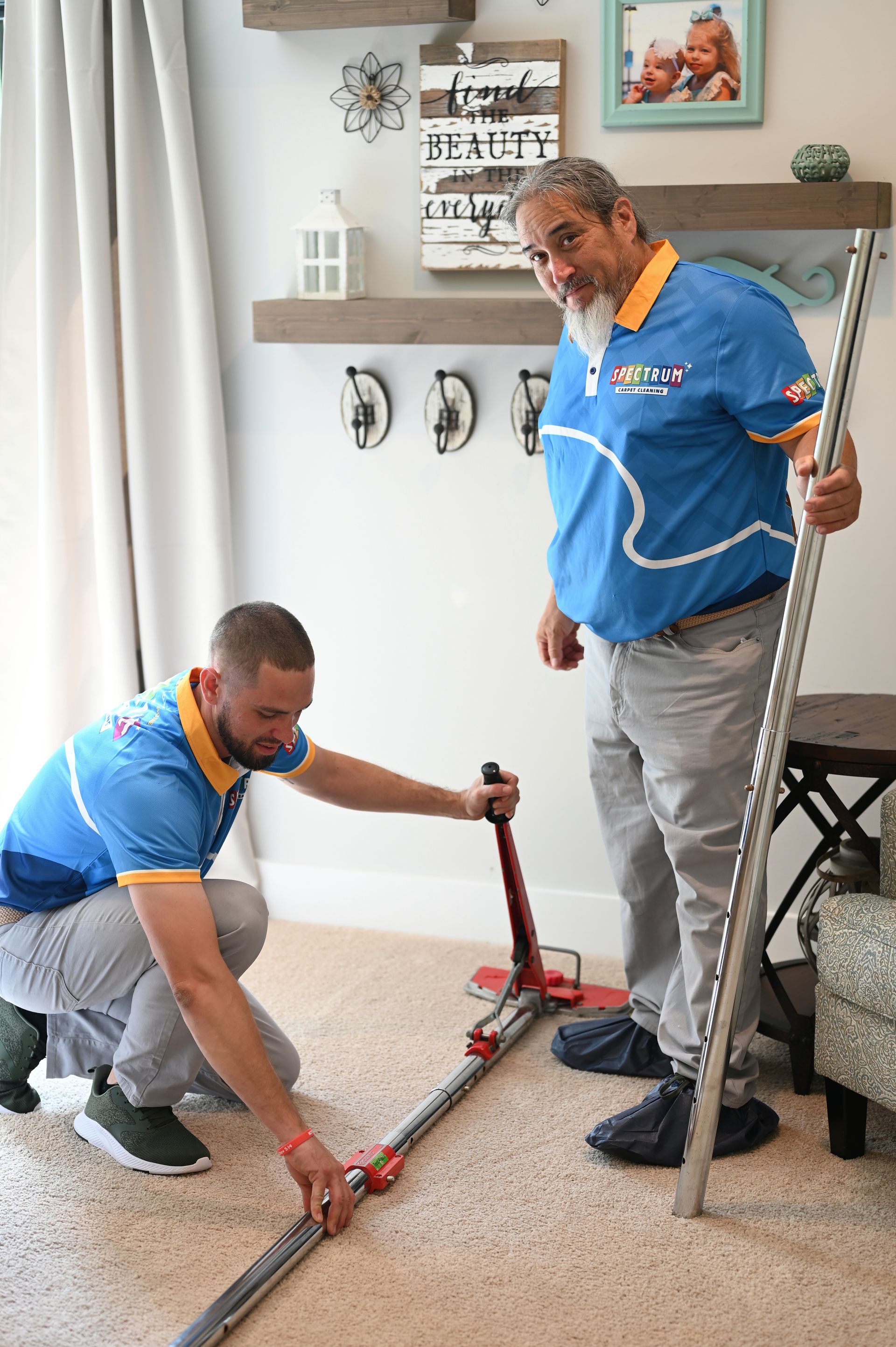 Two men, in blue shirts, installing carpet in a living room. One uses a carpet stretcher.