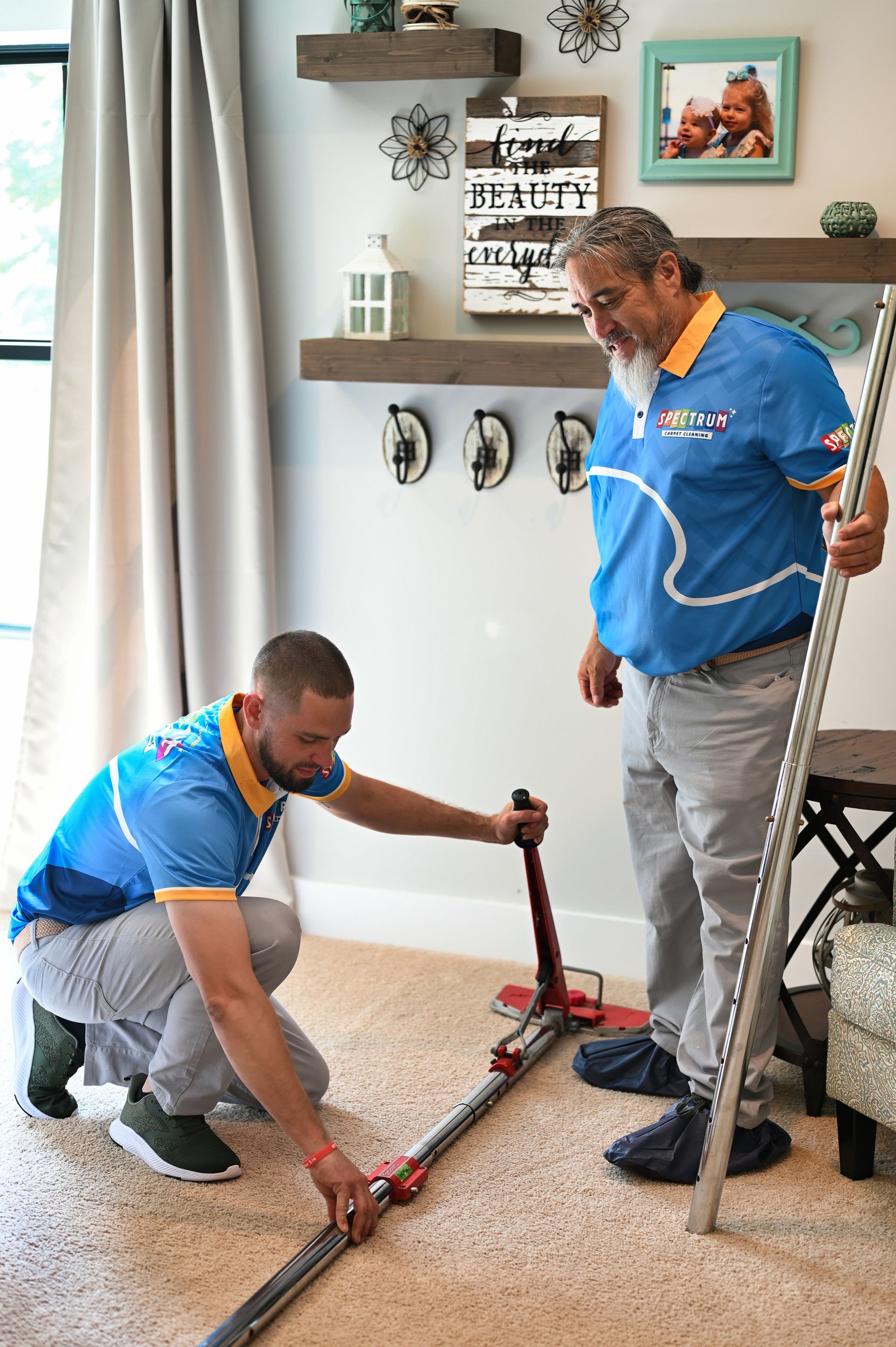 Two men installing carpet in a home. One kneels, operating a tool, the other stands holding a pole.