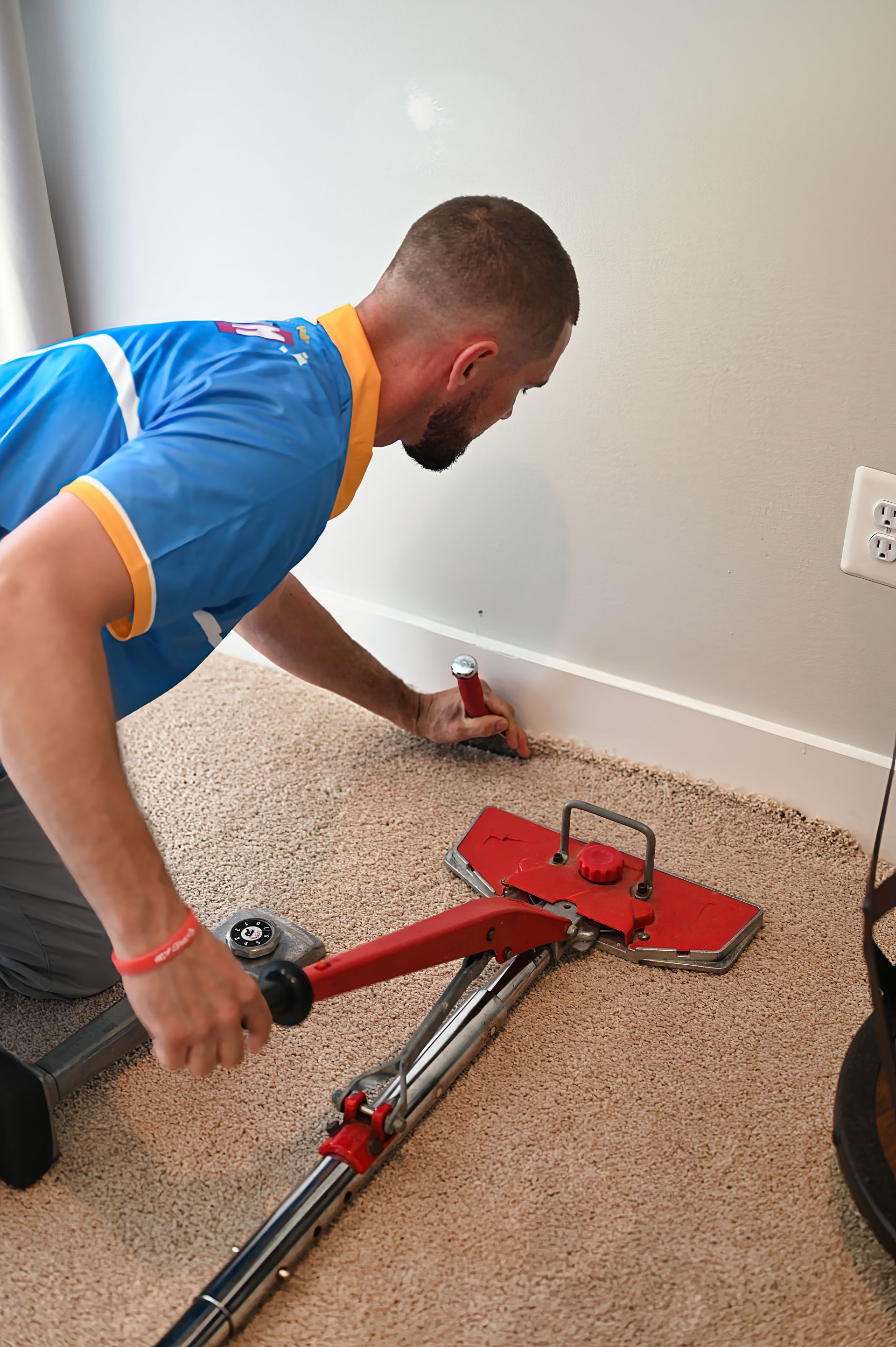 Man installing carpet with a knee kicker and a hammer near a wall; indoors.