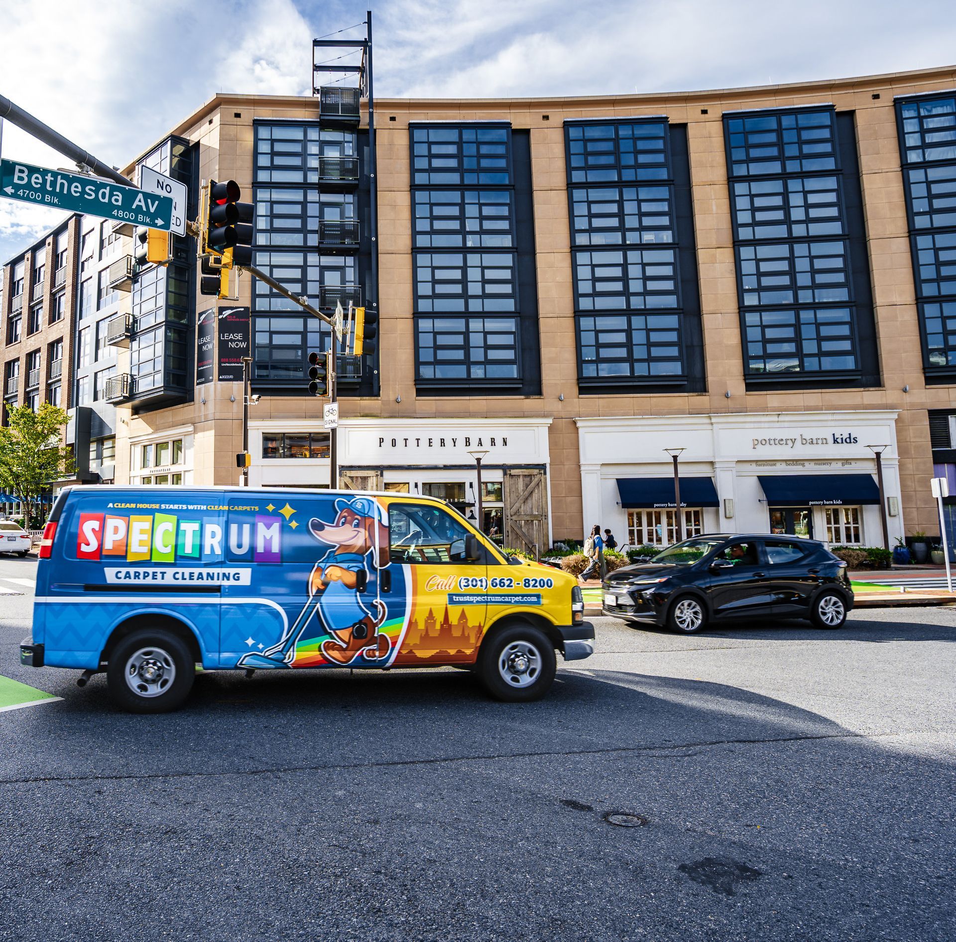 Spectrum van on city street; Bethesda Ave. sign. Tall building, shops, black car, blue sky.