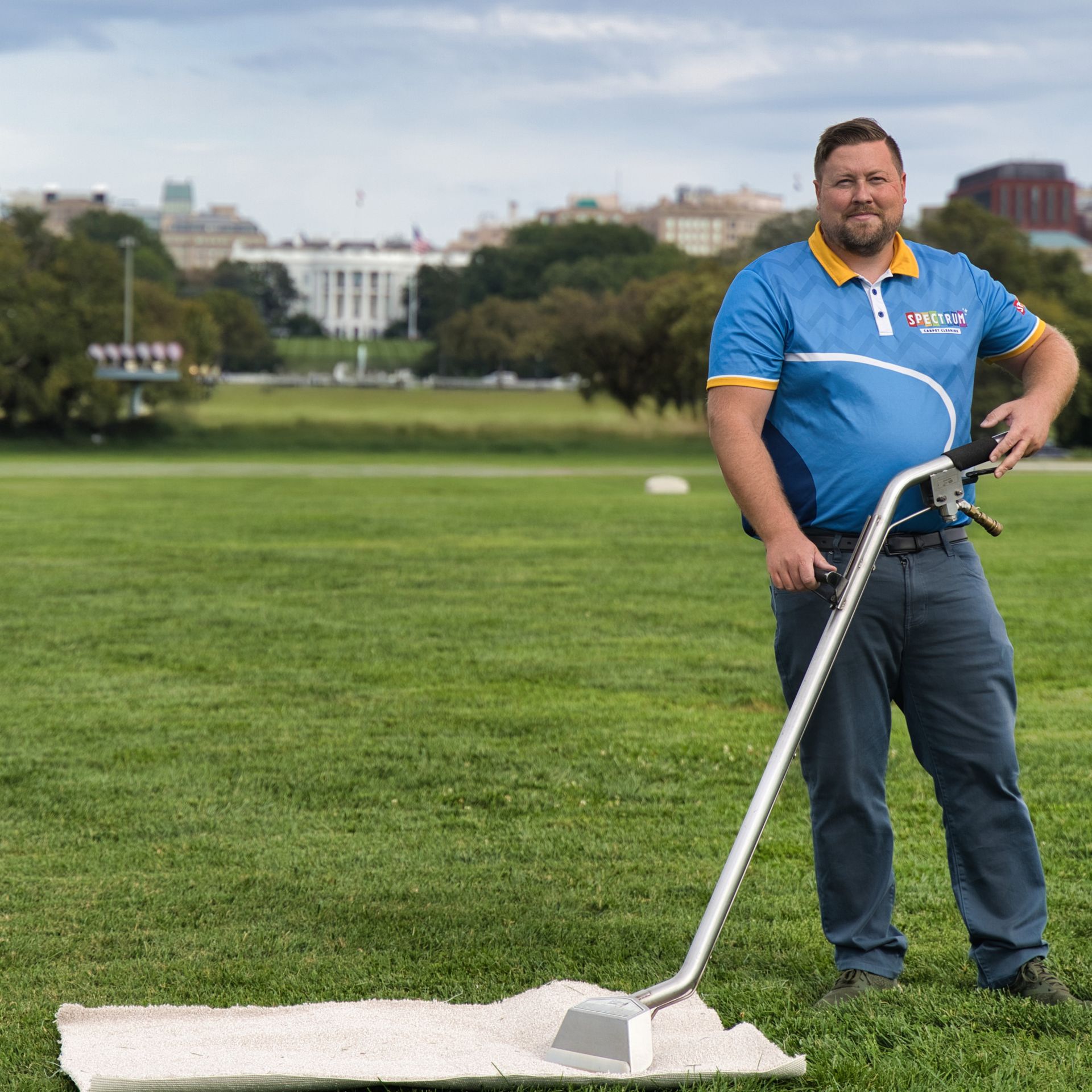 Man in blue shirt vacuums a patch of grass on the National Mall, with the White House in the background.