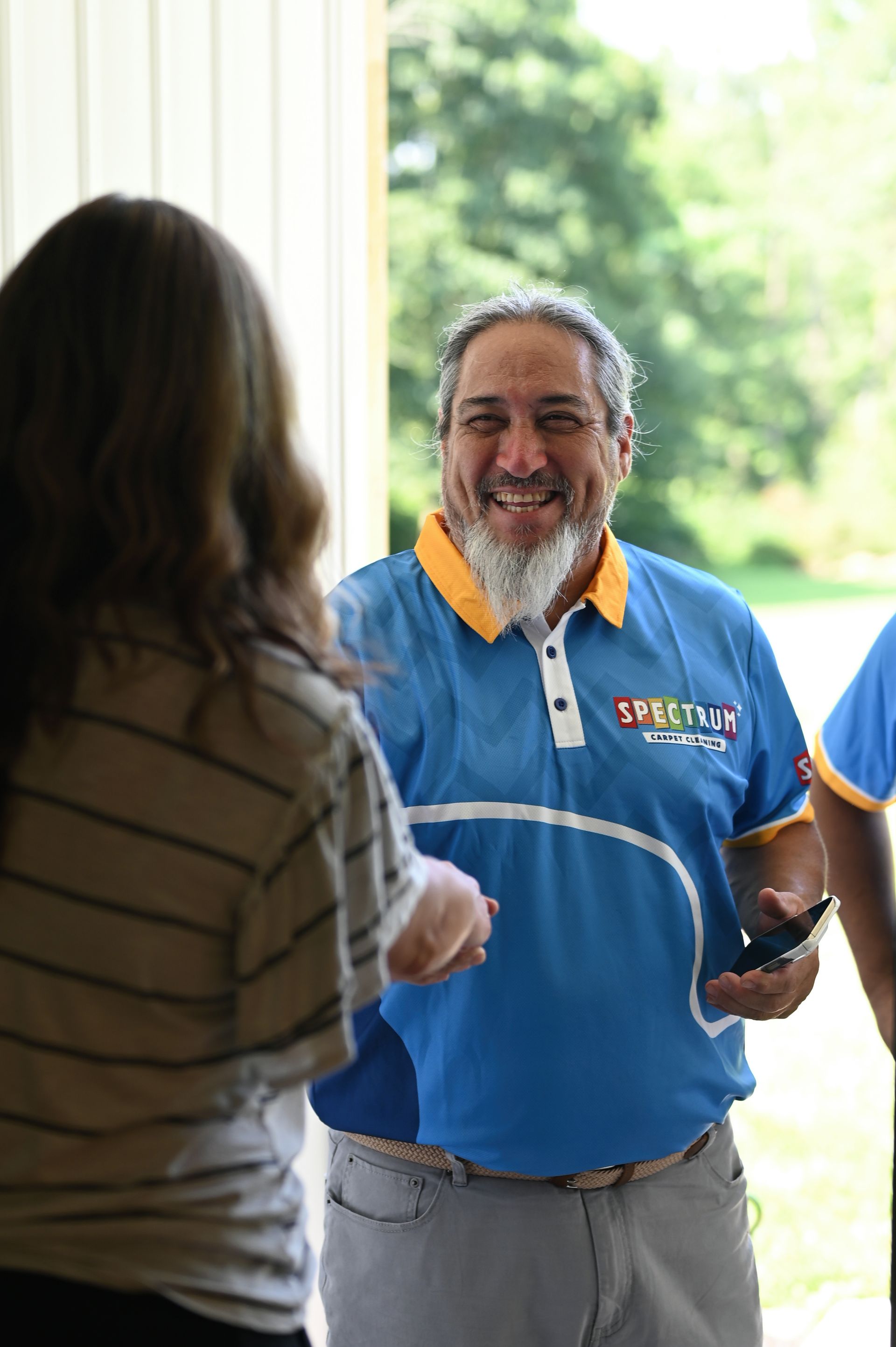 Man in blue Spectrum shirt smiles at a person. Outdoors, daylight.
