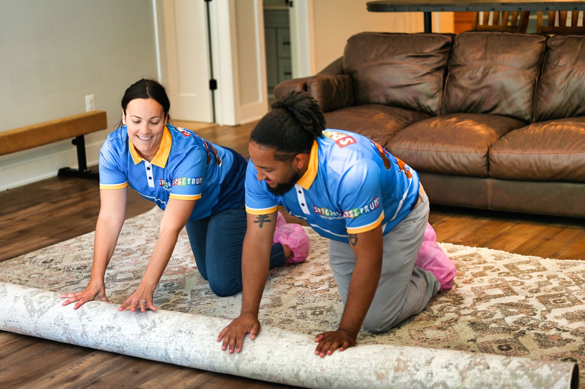 Two people rolling out a patterned rug in a living room. Both are kneeling and smiling, wearing blue shirts.