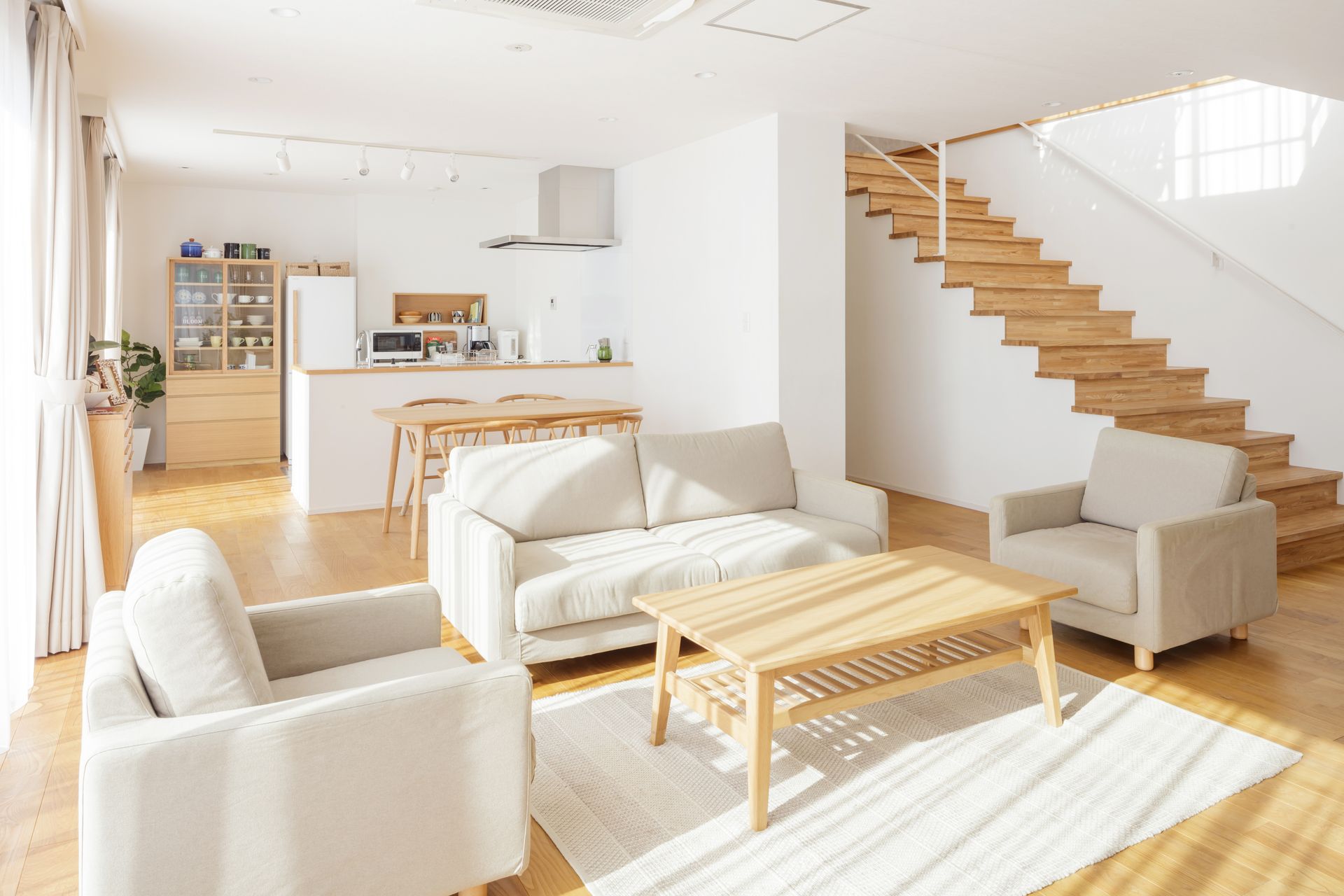 Salon lumineux et aéré avec parquet et escalier en bois clair, canapés blanc cassé et petite cuisine.