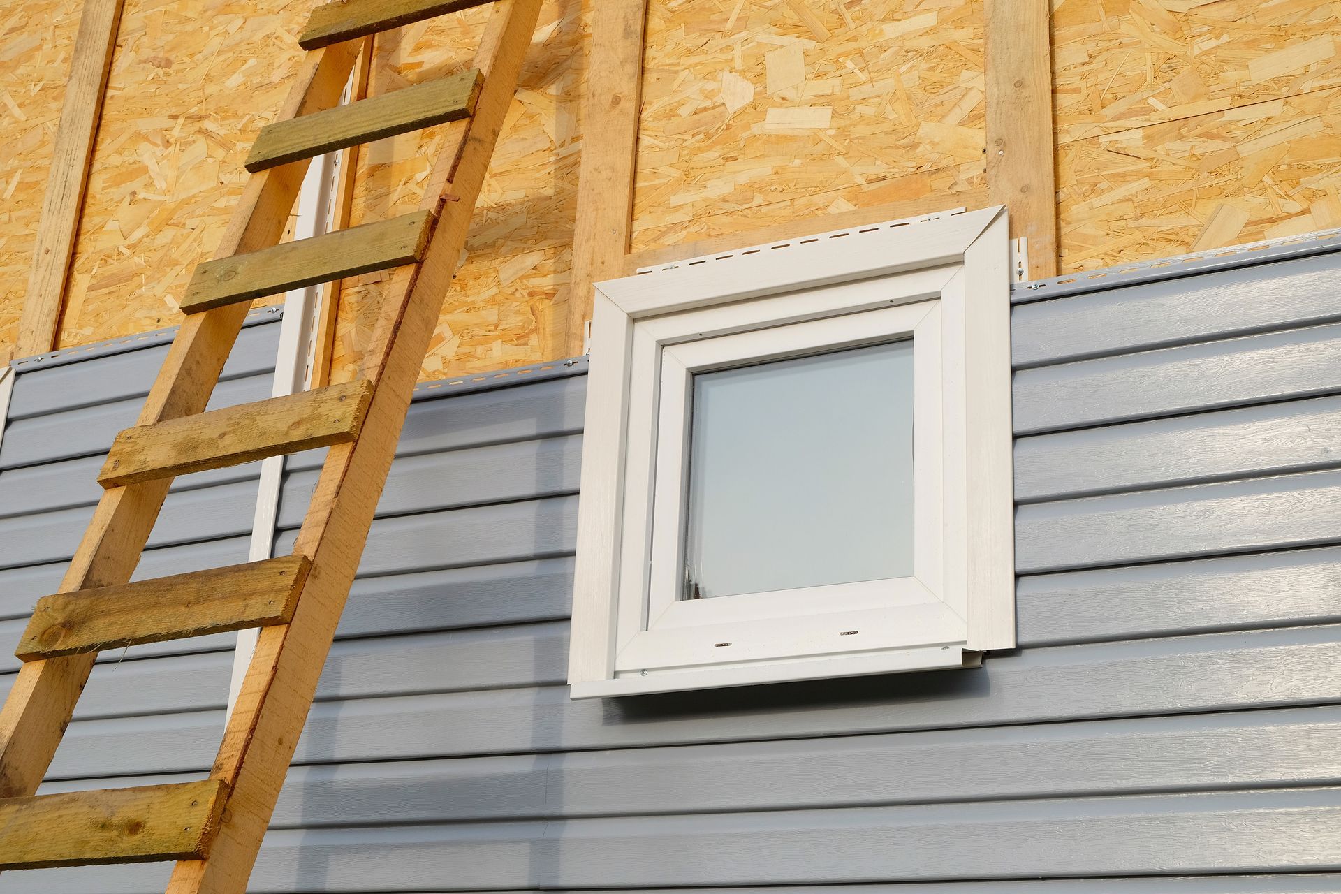 Une échelle en bois est appuyée contre la façade d'un bâtiment dont une fenêtre à cadre blanc est encastrée dans un bardage gris.