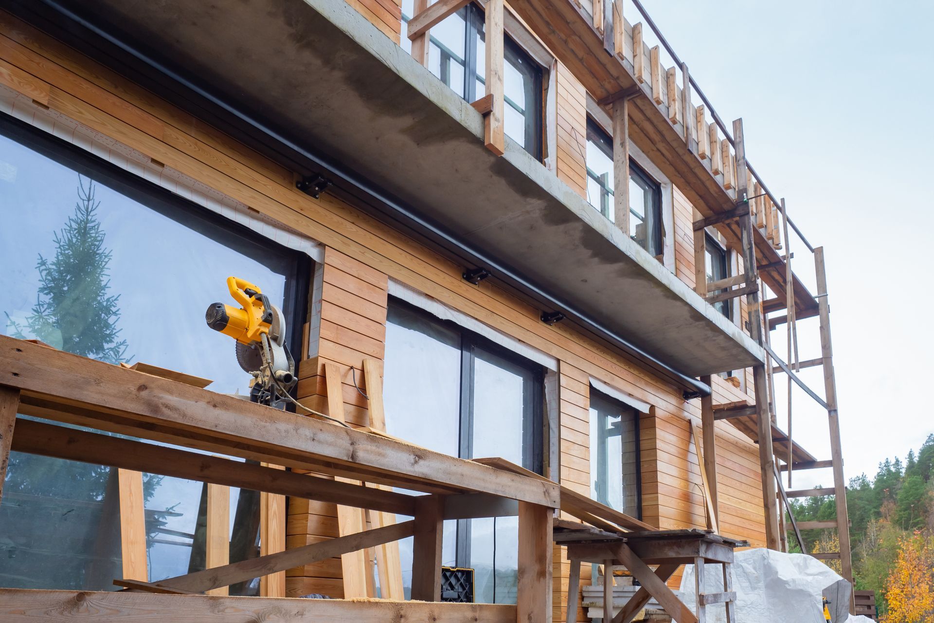 Construction d'un bâtiment à bardage en bois avec de grandes fenêtres ; une scie repose sur un échafaudage.