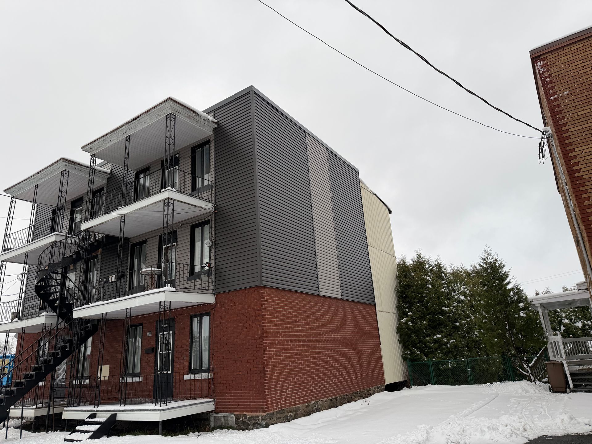 Immeuble d'appartements avec bardage métallique et balcons, soubassement en briques rouges, neige au sol, ciel couvert.
