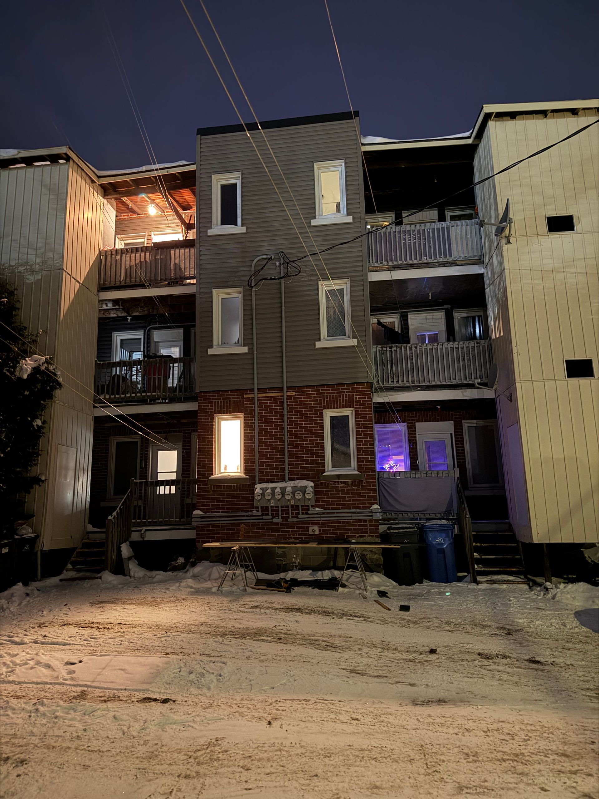 Façade d'un immeuble de trois étages, la nuit. Neige au sol. Plusieurs fenêtres éclairées.