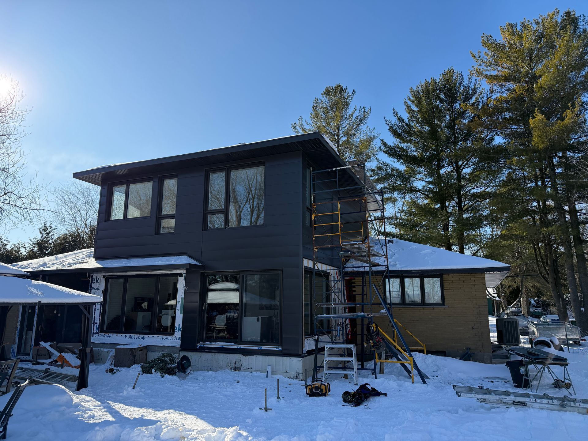 Maison à deux étages en rénovation, au revêtement foncé, entourée de neige et d'arbres. Échafaudages visibles.